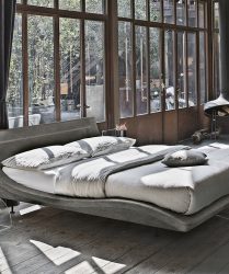 letto moderno di forma ondulata grigio in una camera in stile industriale dominata da colori grigio e marrone con arredo di design e camino sospesp